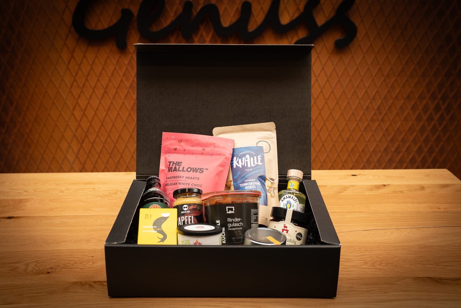 Geöffnete schwarze Geschenkbox, gefüllt mit einer Auswahl an hochwertigen Snacks und Leckereien – darunter Marshmallows, Schokolade und verschiedene verpackte Köstlichkeiten – auf einem Holztisch.