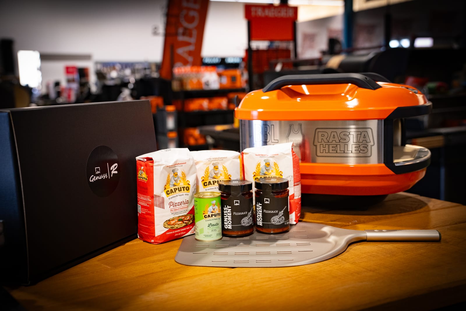Pizza-Geschenkset mit orangefarbenem Pizzaofen, u.a. Pizzamehl, Pizzasauce, Pizzaschieber und Geschenkbox auf einem Holztisch.